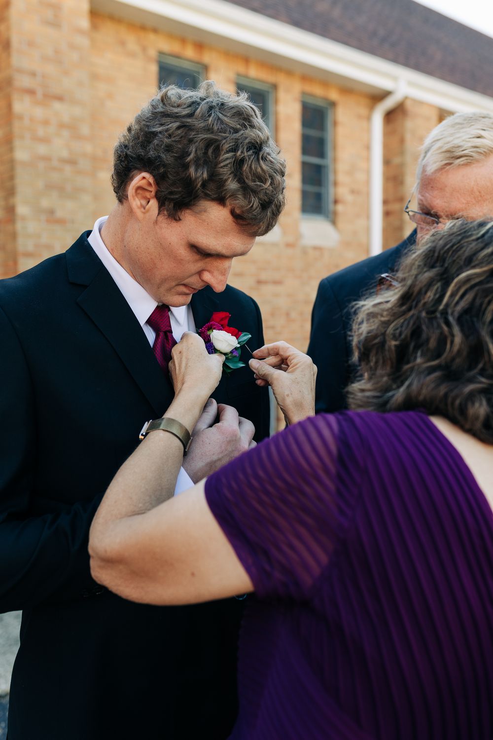 A woman in purple pins a red and white boutonniere on a groom in a black suit outside a brick church.