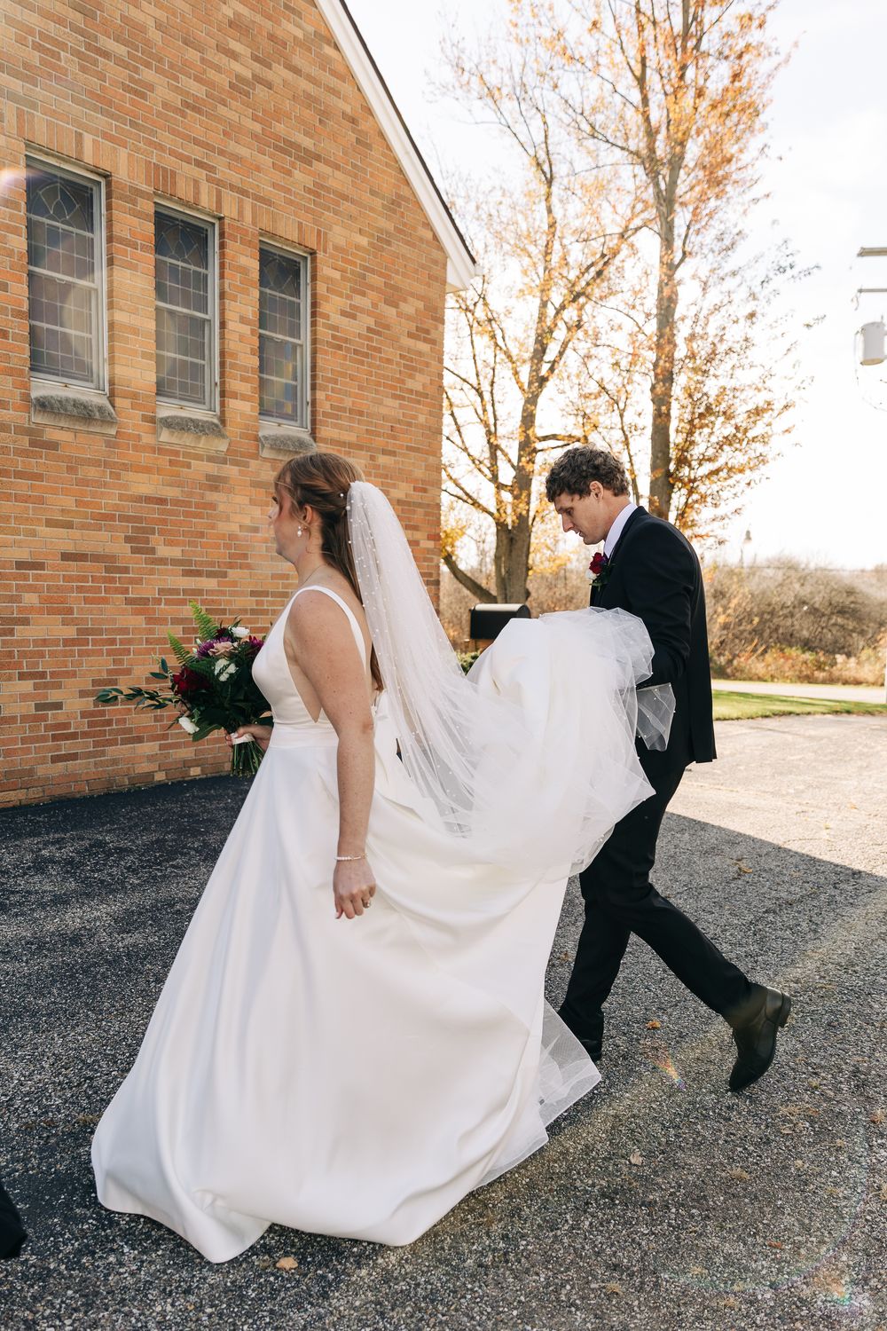 Groom carries bride's white wedding dress train as they walk along a gravel path beside a brick church in autumn.