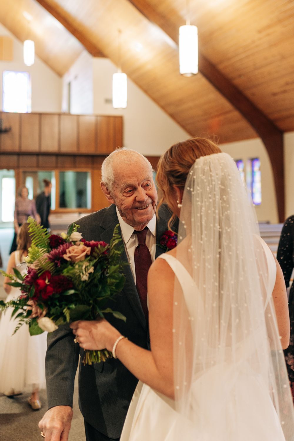 Elderly man smiling warmly at bride holding burgundy bouquet in wooden church aisle.