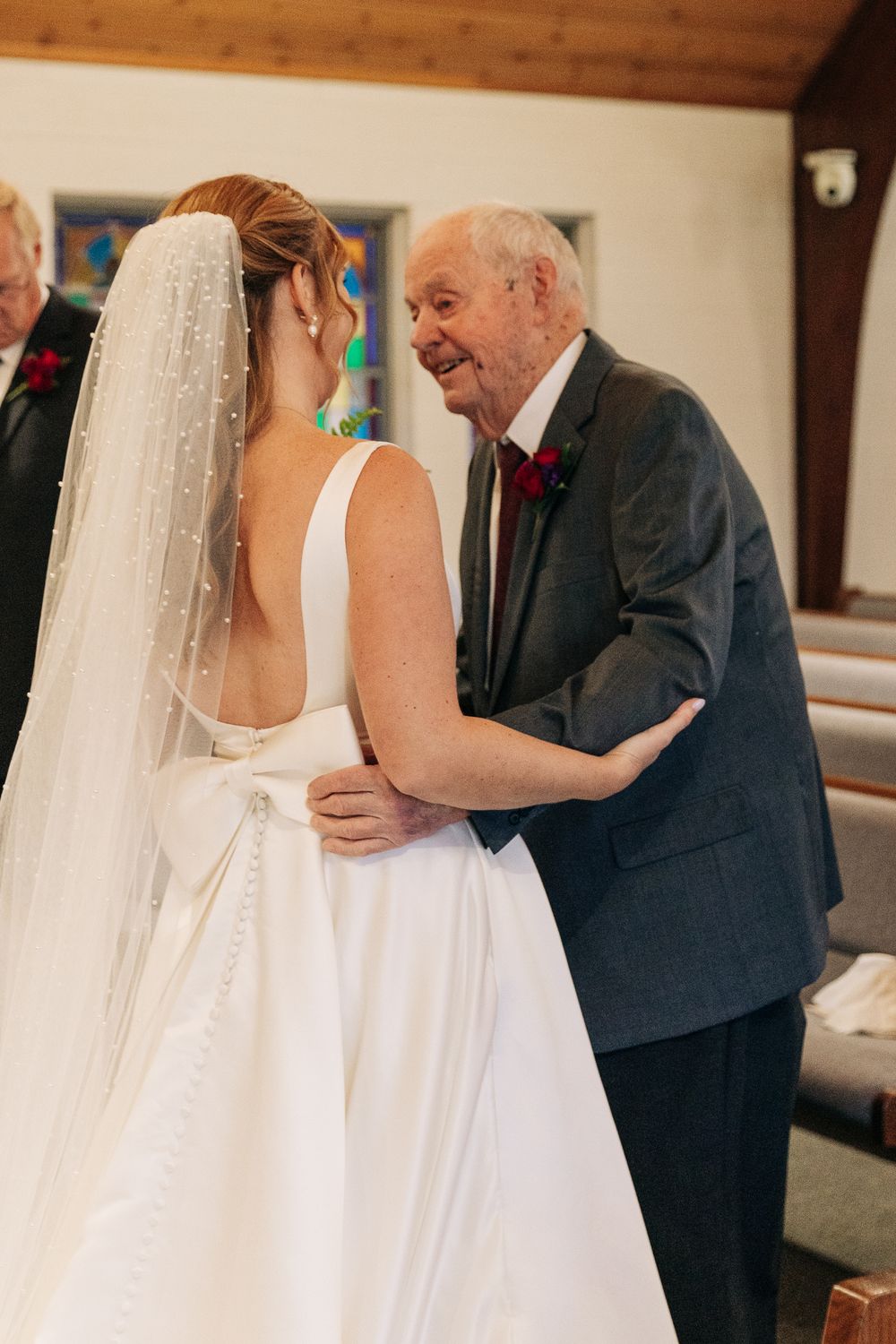 Elderly man in gray suit shares a joyful embrace with bride in white wedding gown and veil inside a church.