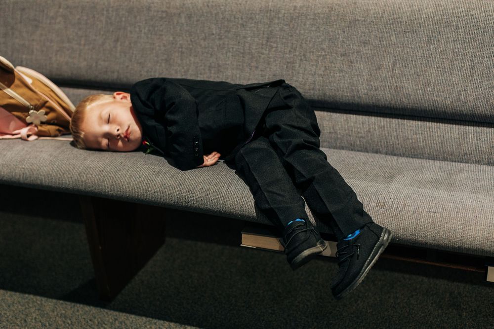 Young child in black outfit sleeping peacefully on a gray upholstered bench.