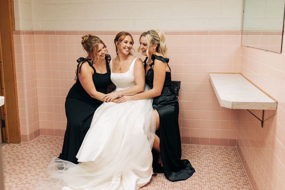 Bride in white gown sits with two bridesmaids in black dresses in a pink-tiled church bathroom before the wedding.