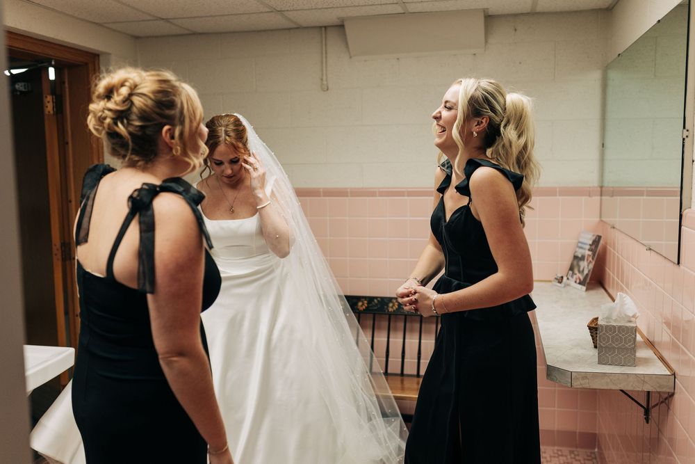 Bride in white gown and veil with bridesmaids in black dresses chatting in a bathroom before the wedding.