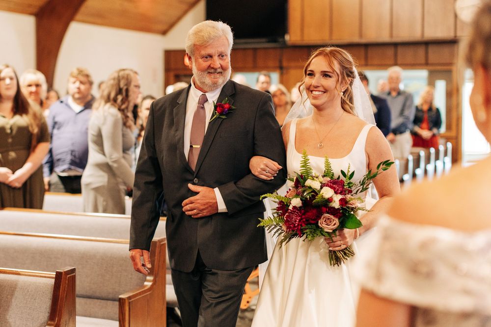 Bride walking down church aisle with father, holding lush red and pink floral bouquet.
