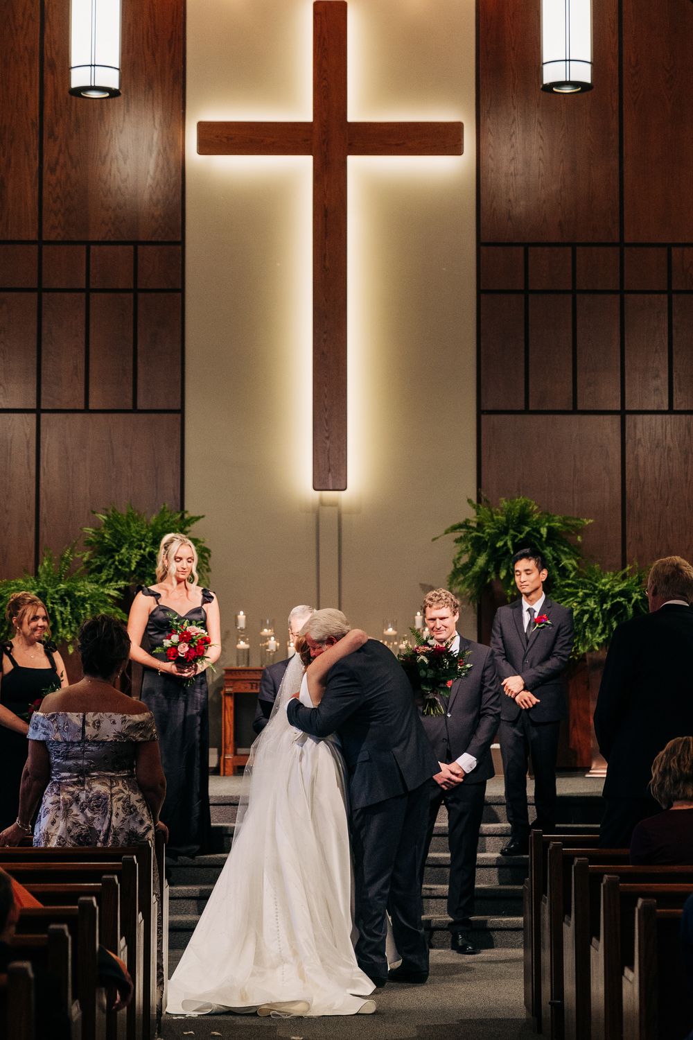 Bride and groom share first kiss at church altar beneath illuminated cross with wedding party and guests watching.