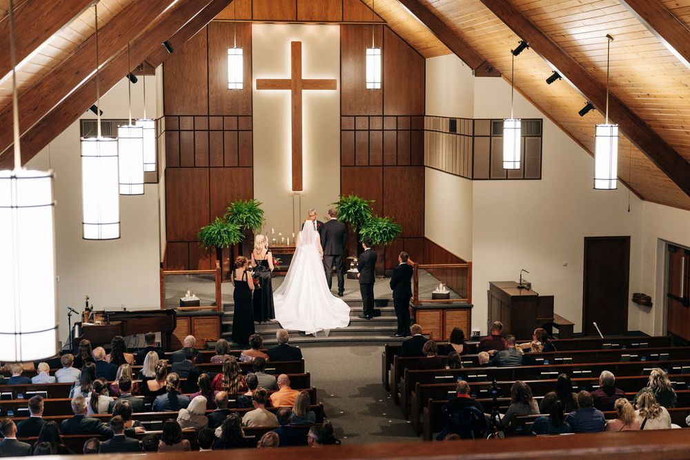 Wide angle view of wedding ceremony at church altar with cross, wedding party, and seated guests in wooden pews.