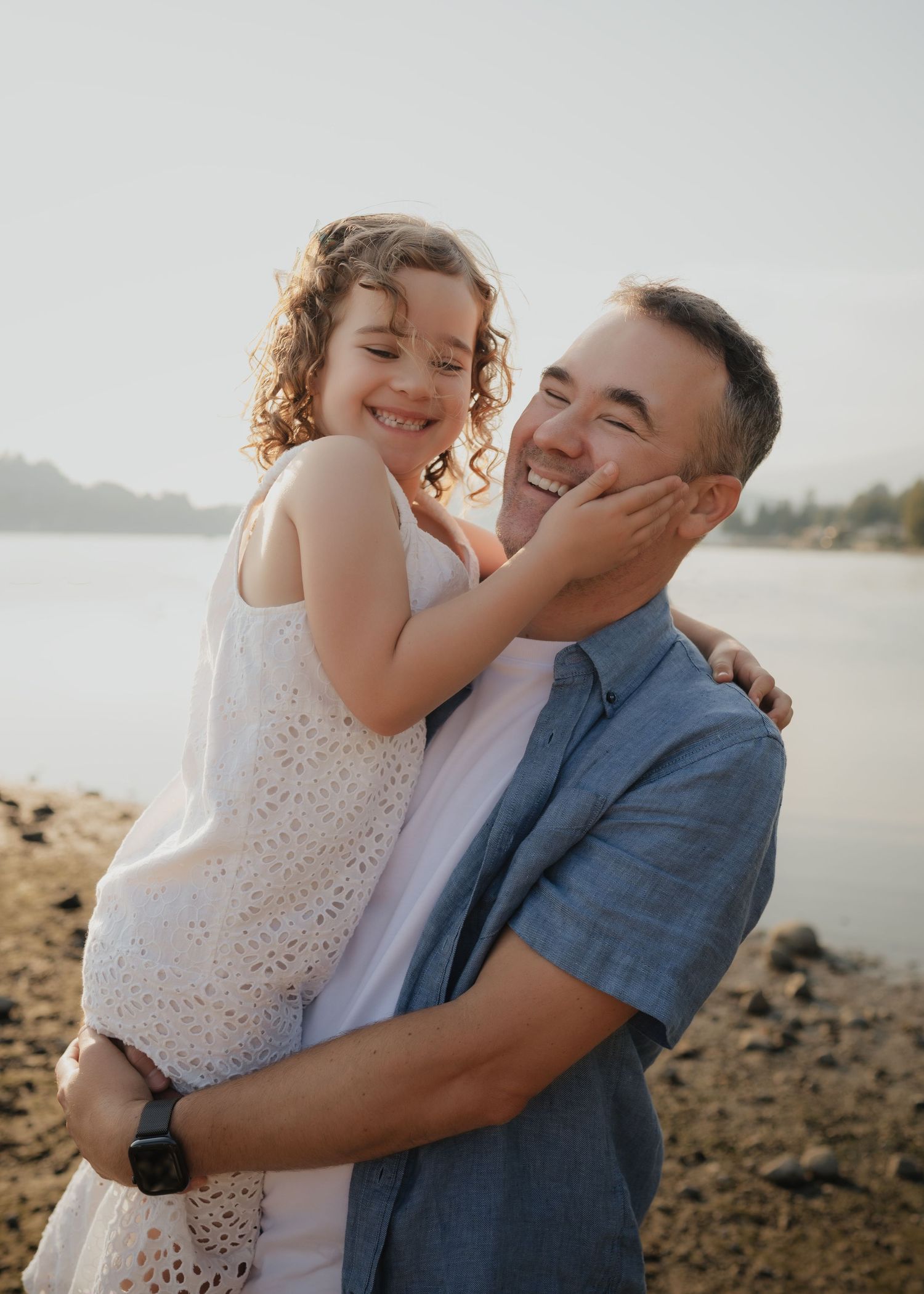A smiling man holds a laughing little girl near a lake shore during golden hour.