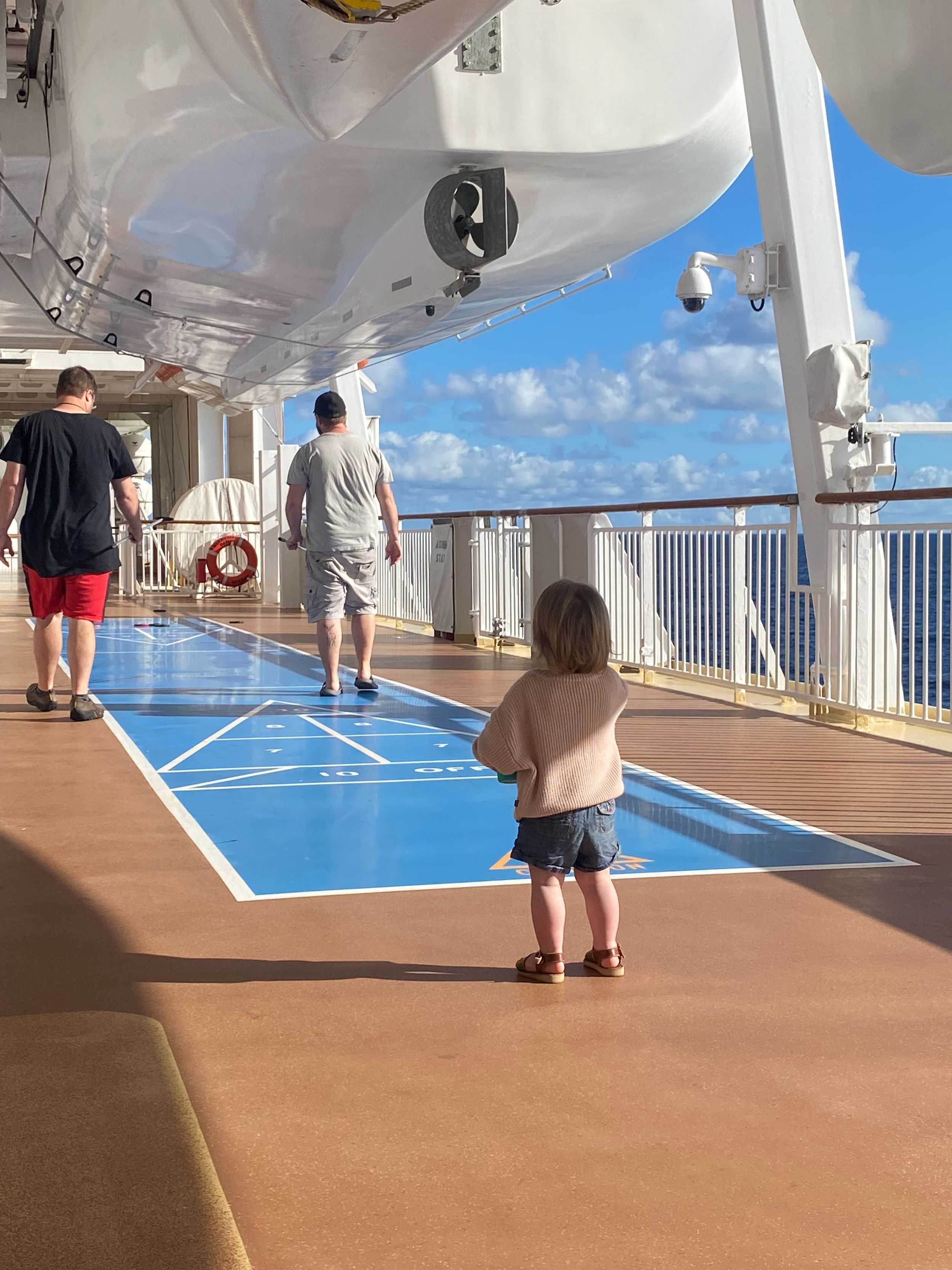 Toddler standing on cruise ship deck near shuffleboard court with adults walking ahead and ocean views.