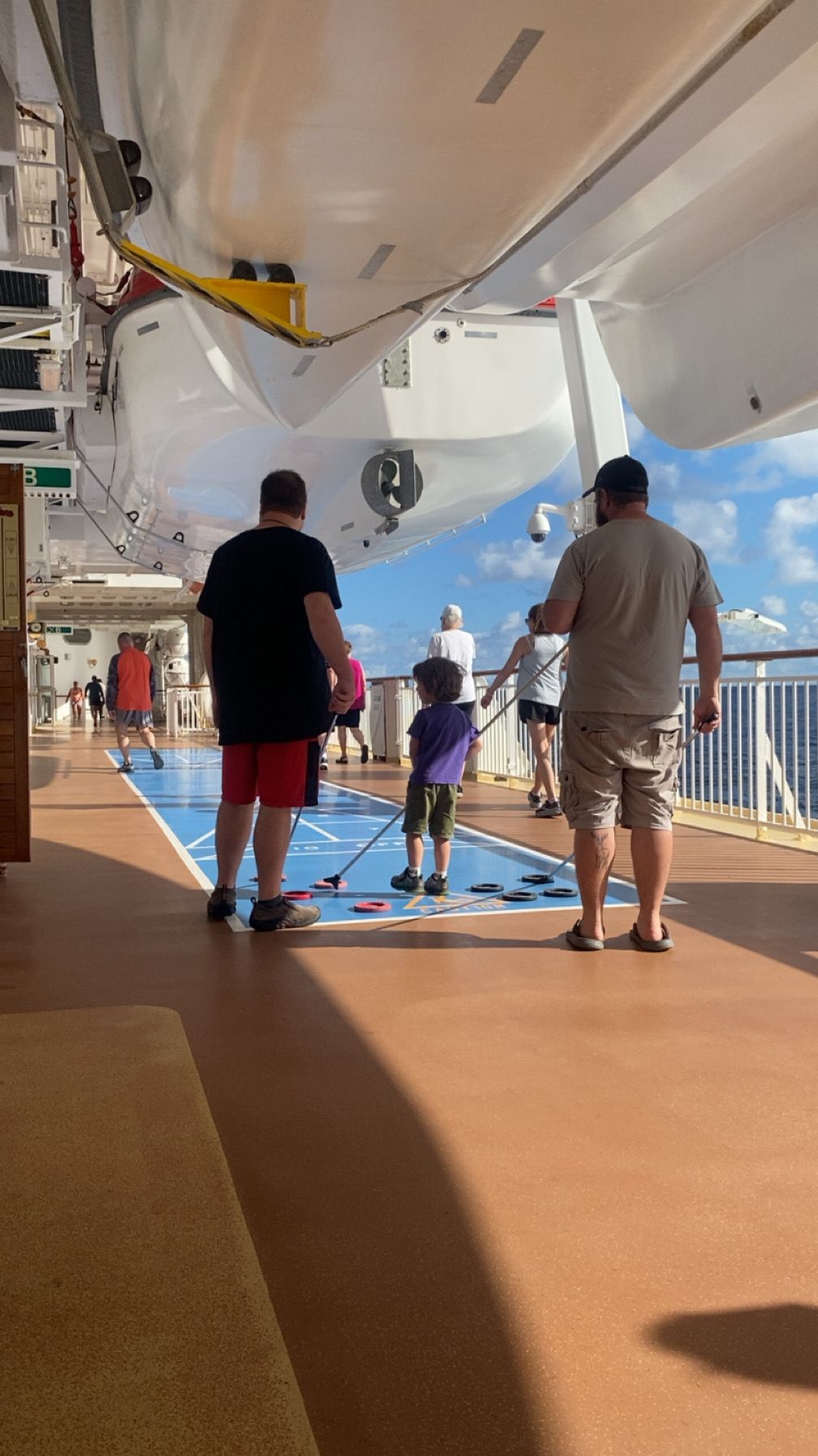 Families playing shuffleboard on a cruise ship deck under a sunny blue sky.