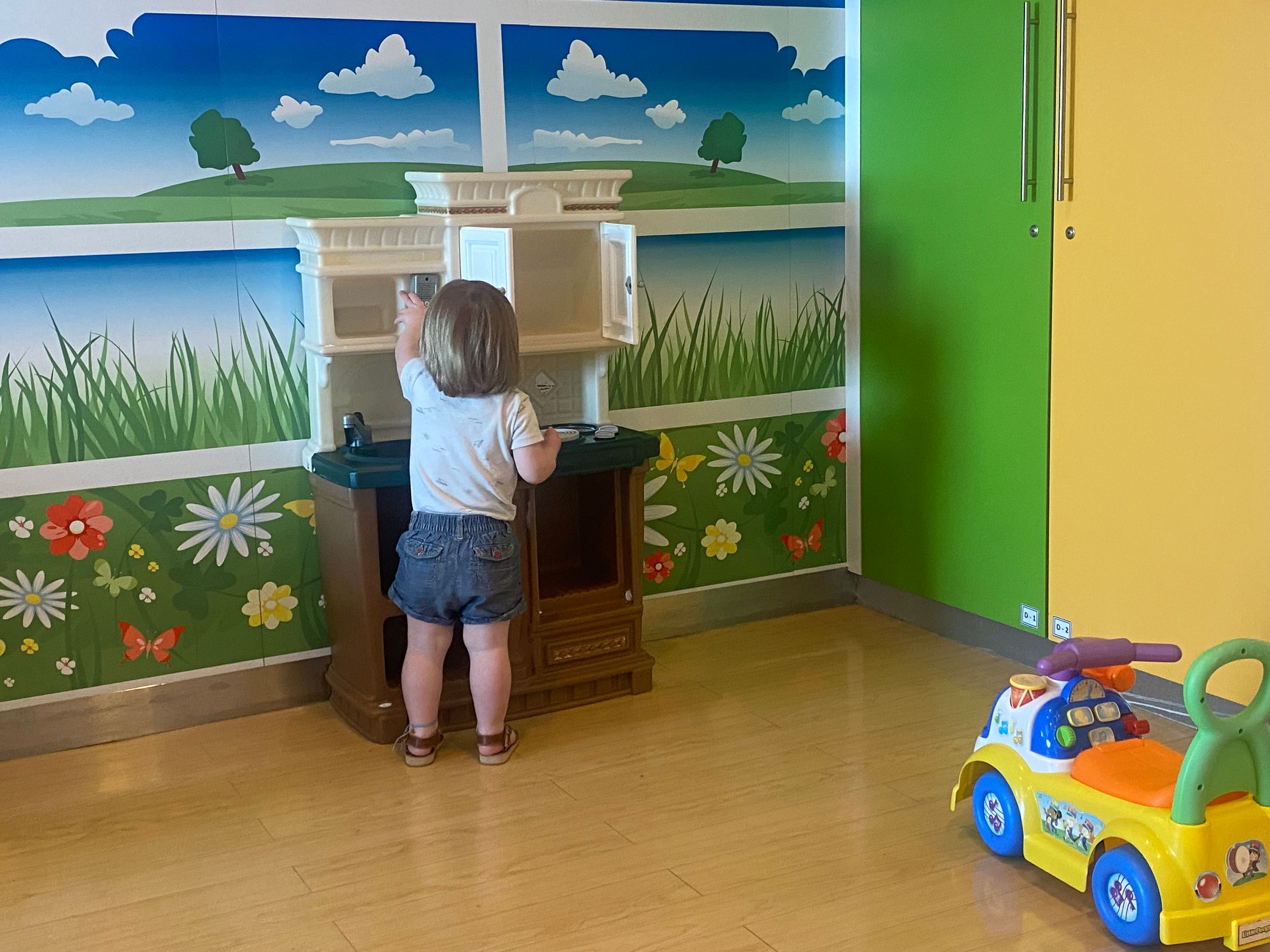 Toddler playing at a wooden toy piano in a colorful children's playroom with nature mural and ride-on toy car.