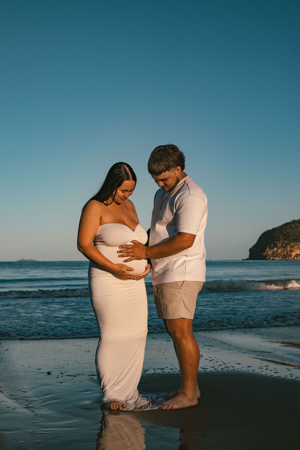 Expectant couple tenderly cradling baby bump on beach at golden hour with ocean waves.