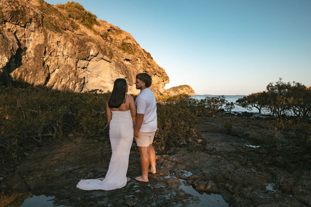 Couple in wedding attire standing on rocky coastal cliff at sunset with ocean views.