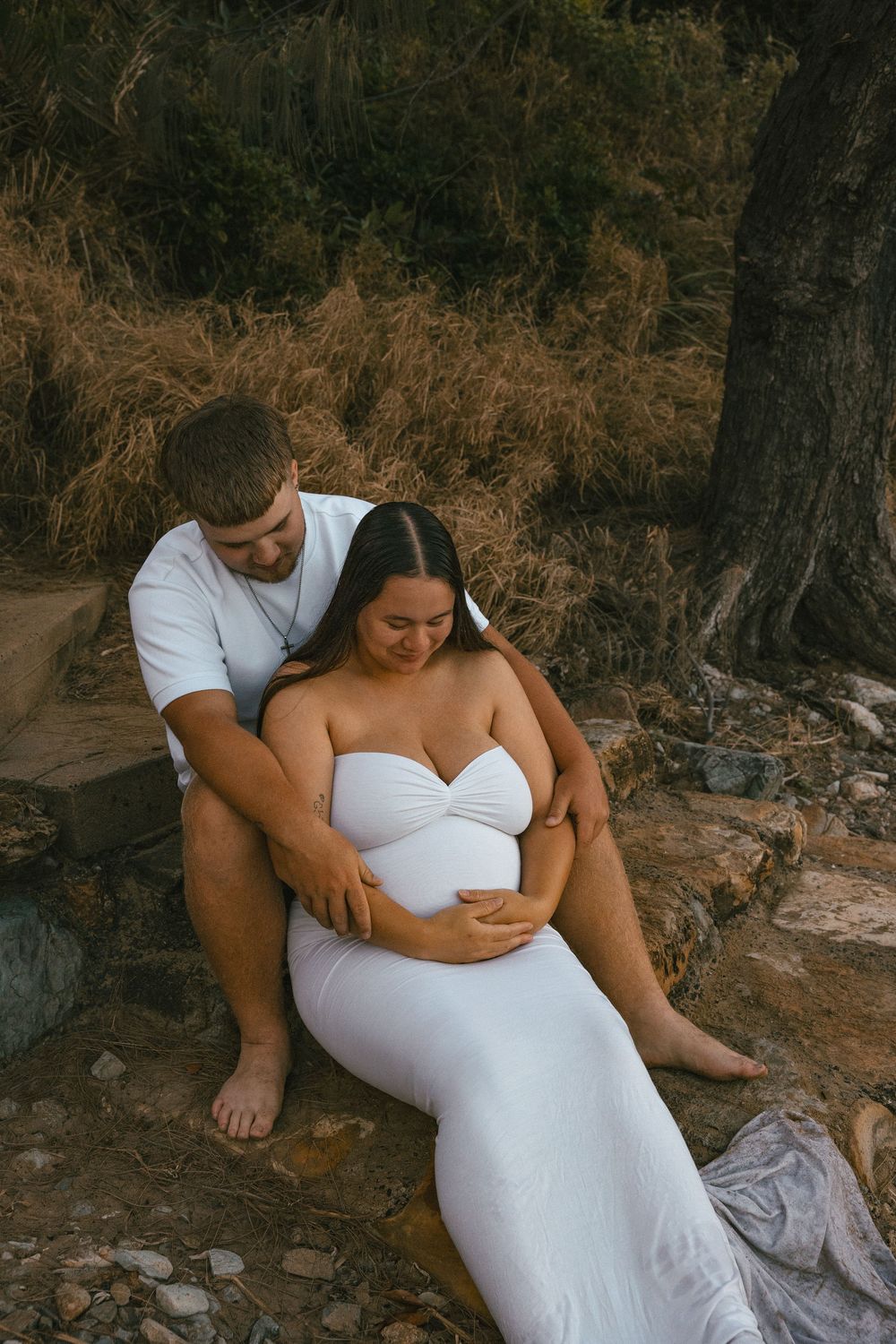 Pregnant couple sitting peacefully outdoors, man embracing woman in white maternity dress.