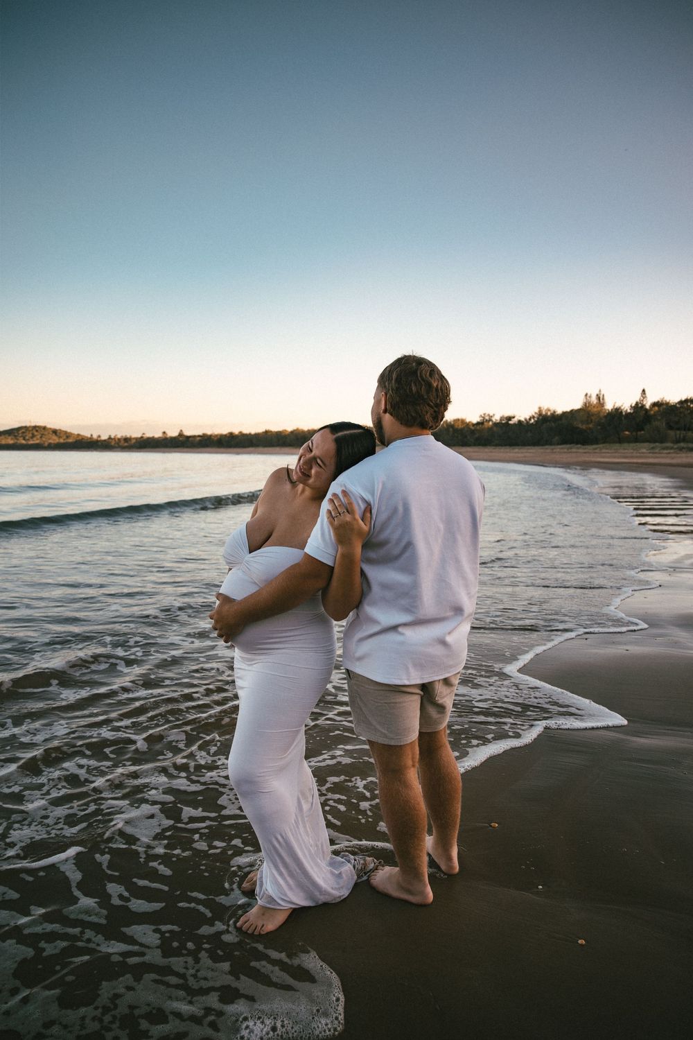 Pregnant couple embracing on beach at sunset, woman in white maternity dress holding baby bump.