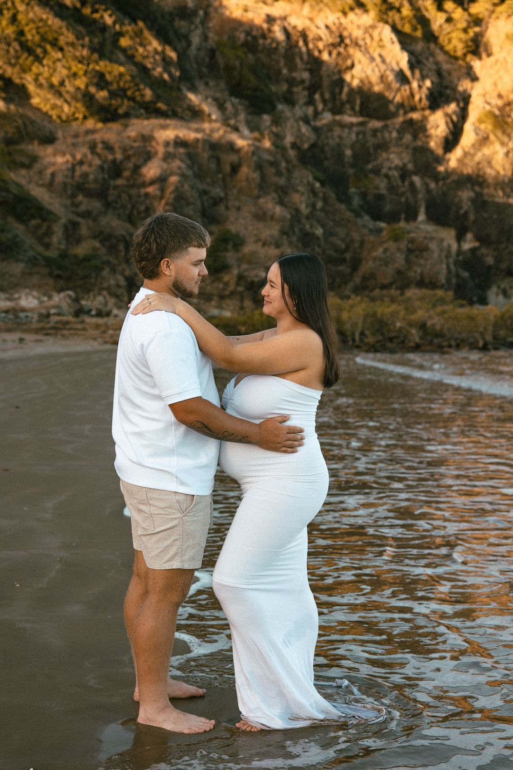 Pregnant couple standing barefoot in ocean waves at golden hour, woman in white dress, man in white shirt and khaki shorts.