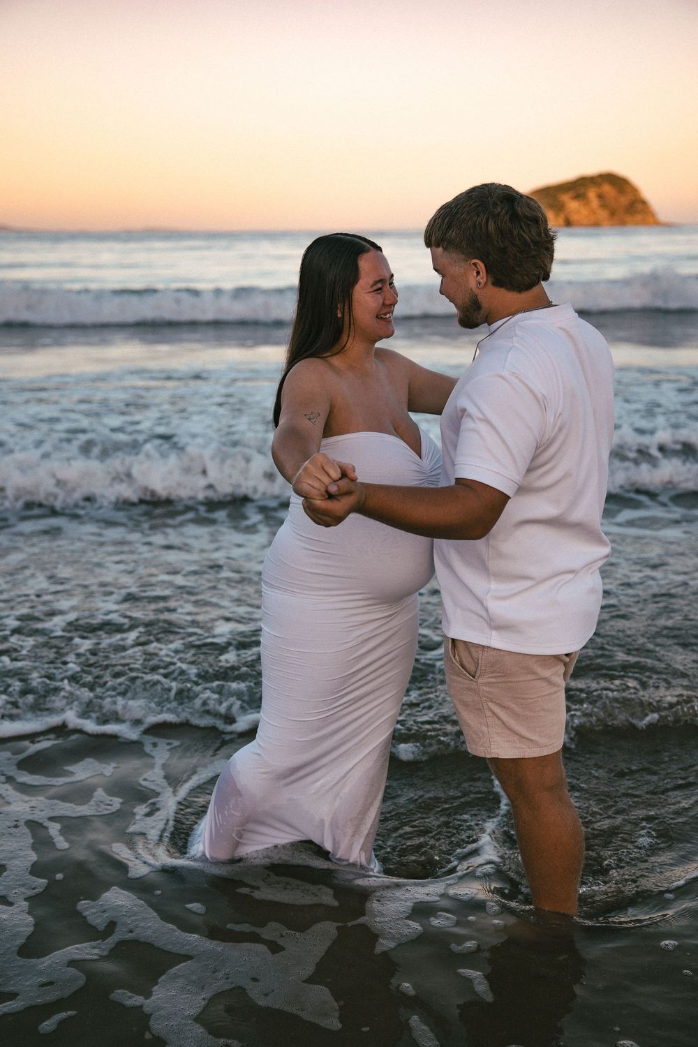 Pregnant couple standing in ocean waves at sunset, woman in white dress, man in white shirt and khaki shorts.