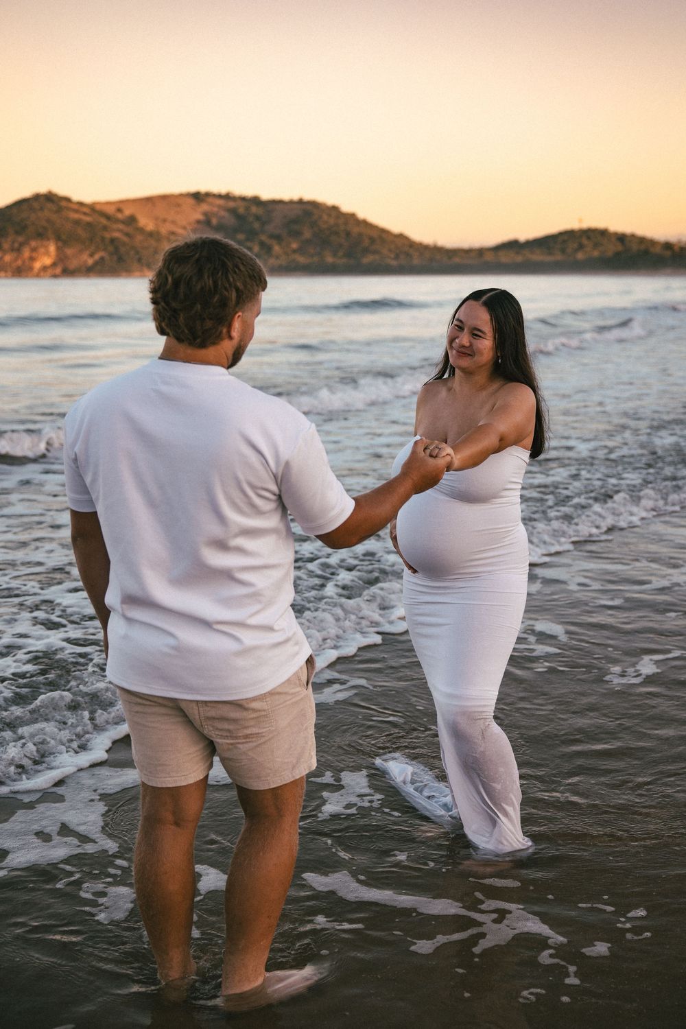 Pregnant woman in wet white dress holds hands with partner in ocean waves at sunset.