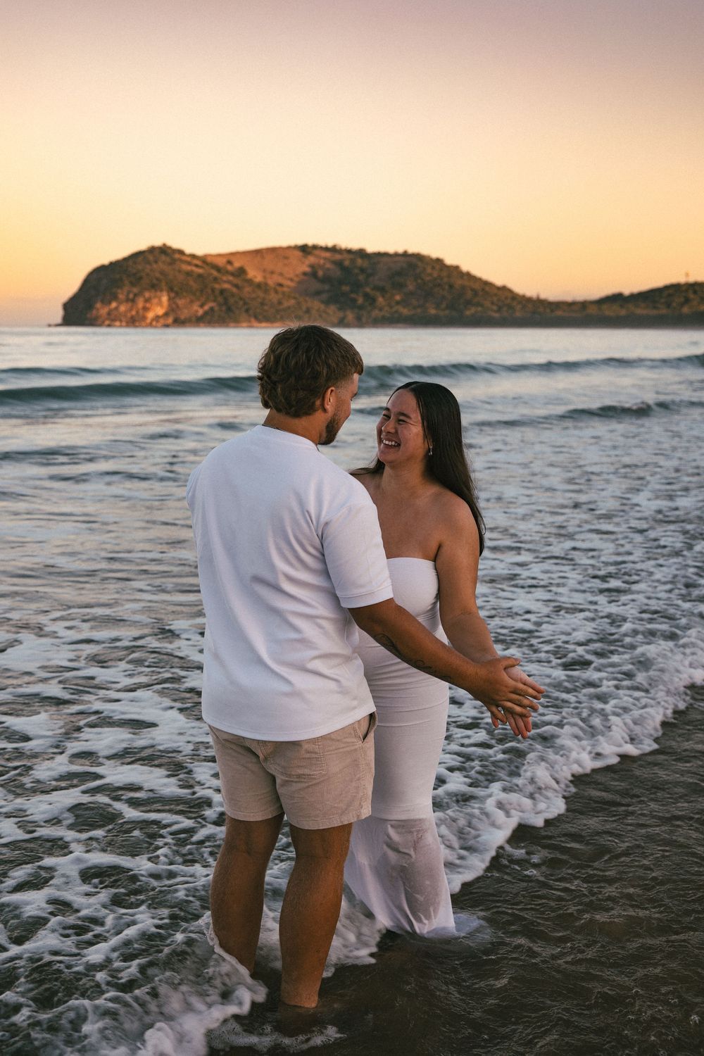Happy couple laughing together in ocean waves at sunset, woman in white strapless maternity dress.