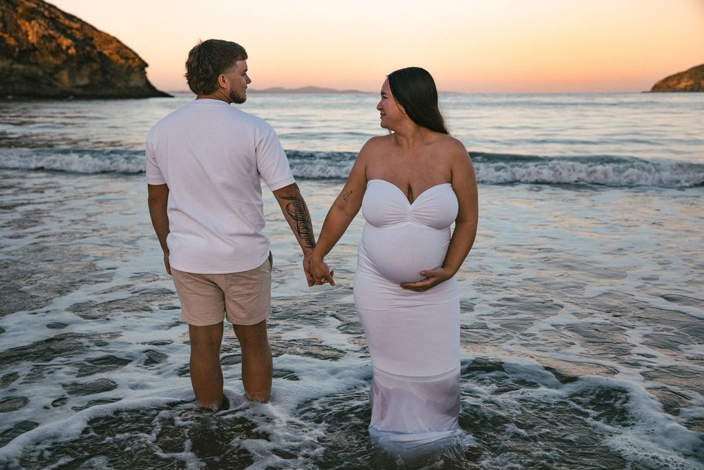 Pregnant couple holding hands at sunset beach, woman in white maternity gown cradling baby bump.