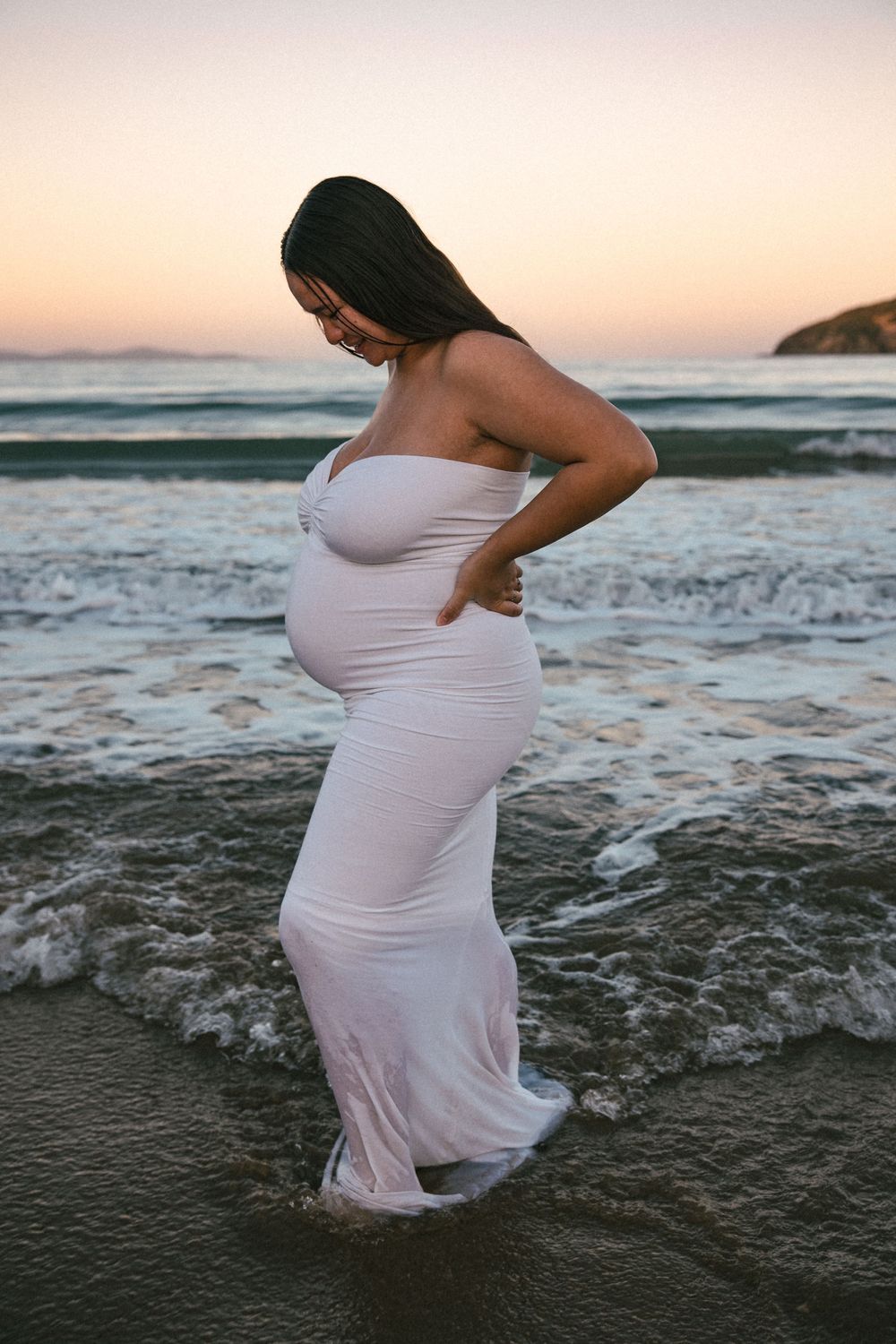 Pregnant woman in white strapless dress standing in ocean waves at sunset, holding her baby bump.