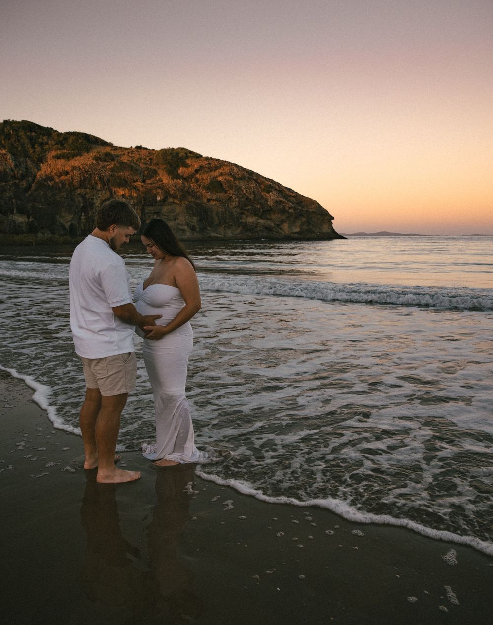 Expecting couple standing in beach waves at sunset, lovingly gazing at pregnant belly.