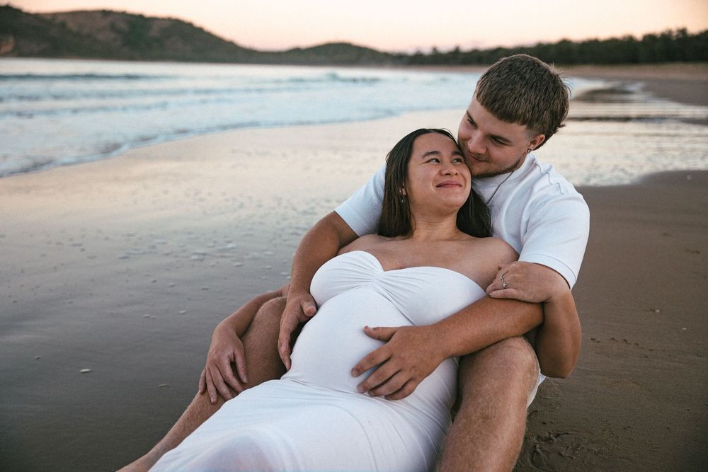 Couple sharing tender beach maternity moment at sunset, woman in white dress leaning against partner.