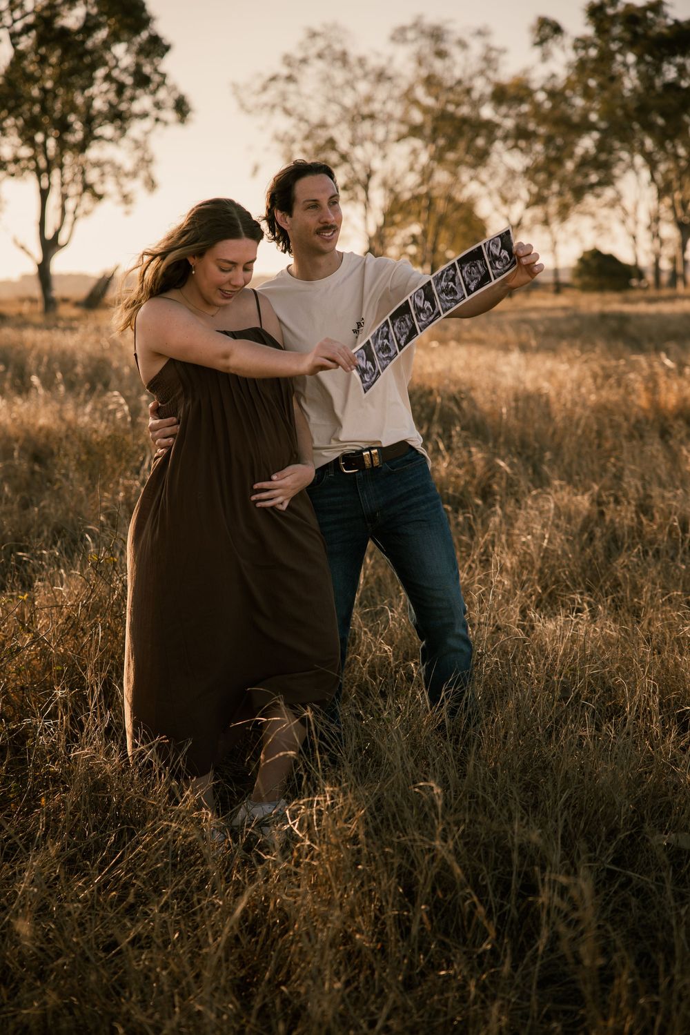 Couple holding ultrasound scans in golden field at sunset, pregnant woman in dark dress embraced by partner. Rockhampton Queensland