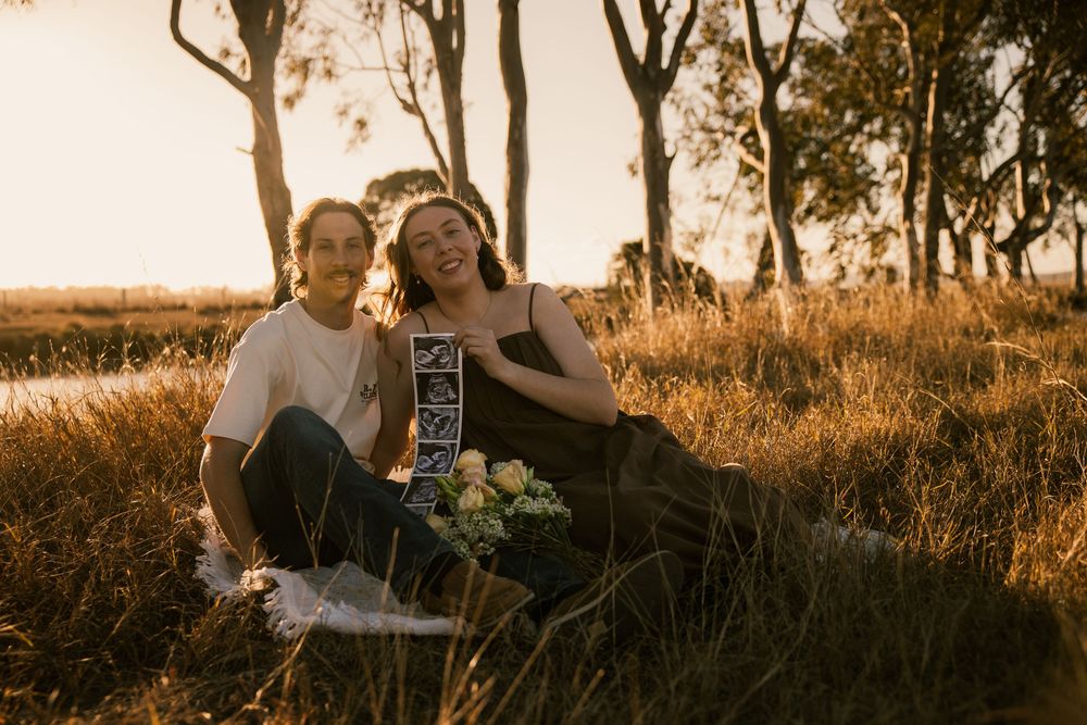 Couple sitting in golden grass at sunset holding ultrasound photos, surrounded by trees in a warm outdoor setting. Rockhampton Queensland