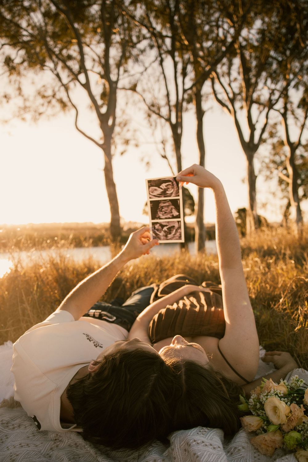 Couple lying in golden field holding ultrasound scan photos up, pregnancy announcement at sunset. Rockhampton Queensland