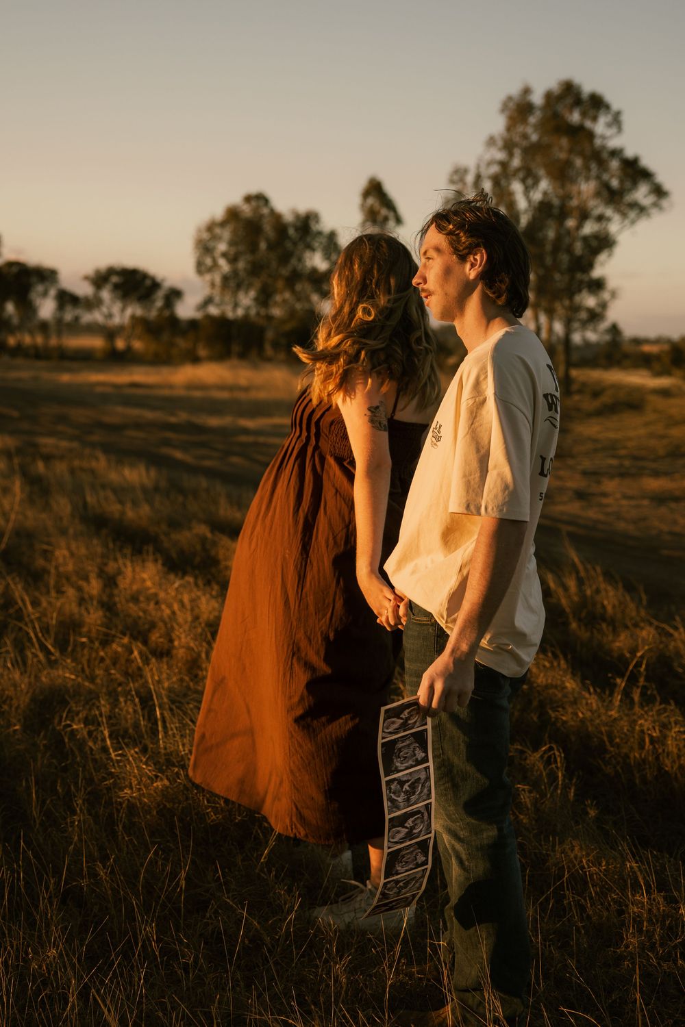 Couple holding hands in golden field at sunset, man holding ultrasound photos, maternity announcement outdoors. Rockhampton Queensland