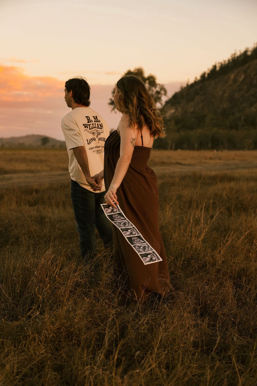 Couple holding hands in golden field at sunset, woman in brown dress holding ultrasound photos, announcing pregnancy. Rockhampton queensland