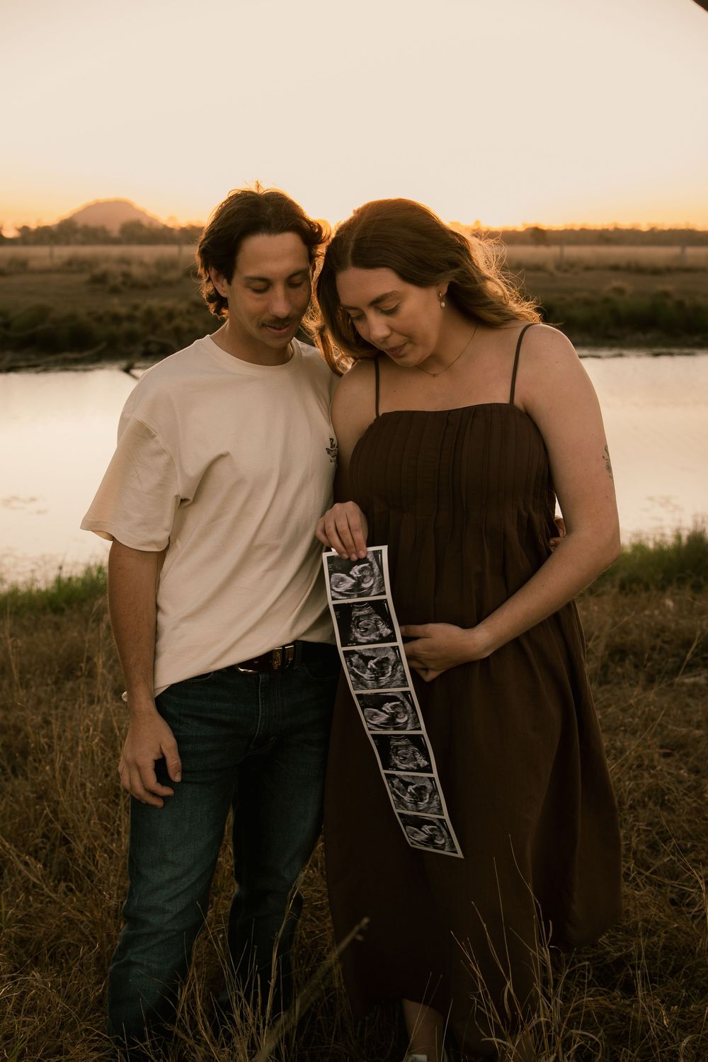 Expectant couple holding ultrasound strip at sunset near a river, announcing their pregnancy outdoors. Rockhampton Queensland maternity