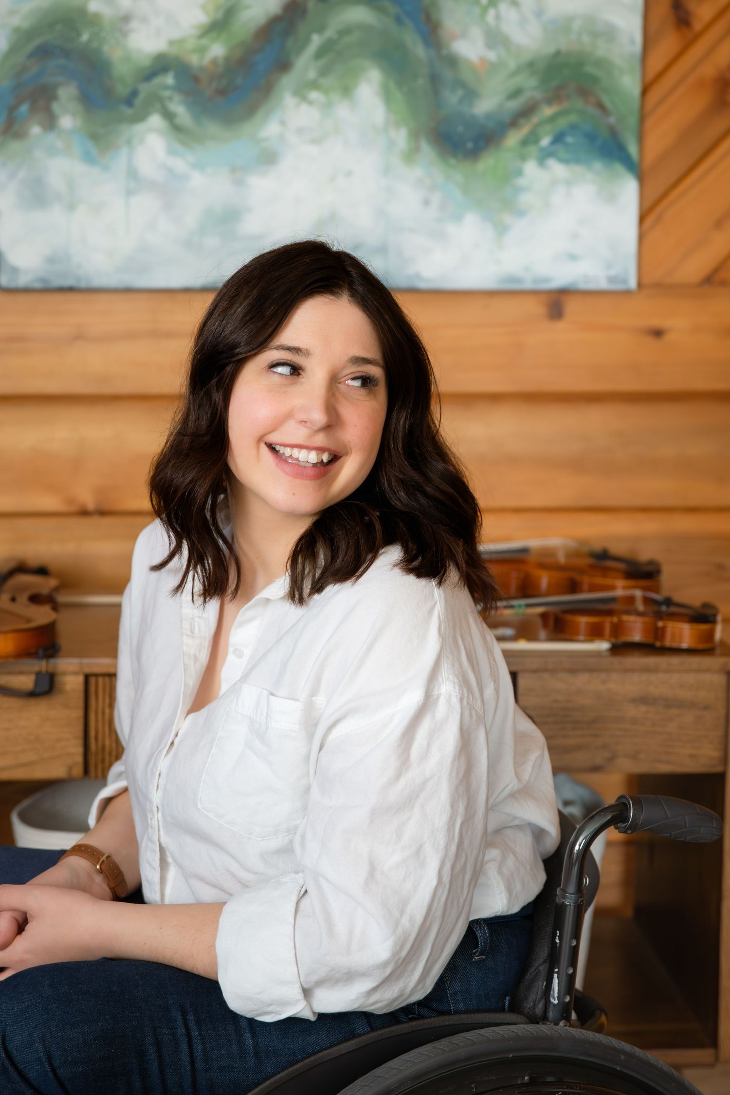 Smiling young woman in a wheelchair wearing a white shirt, seated in a cozy wood-paneled room with violins and abstract art.