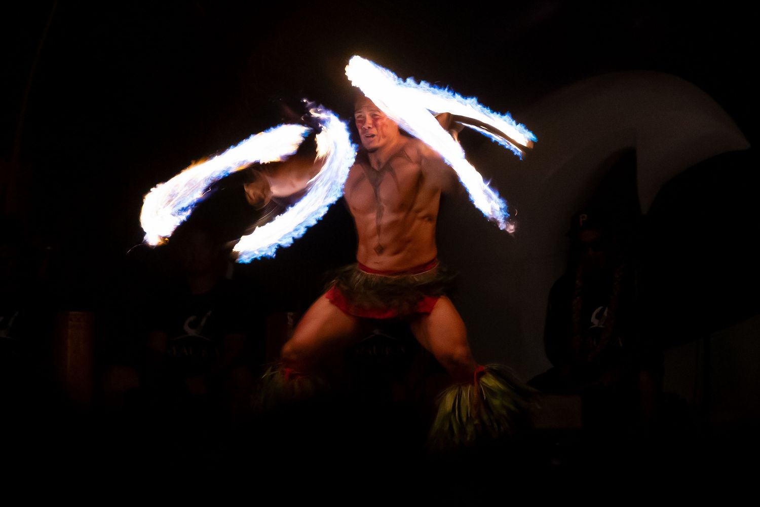 Fire dancer performing at night, holding flaming torches with arms outstretched, wearing traditional grass skirt.