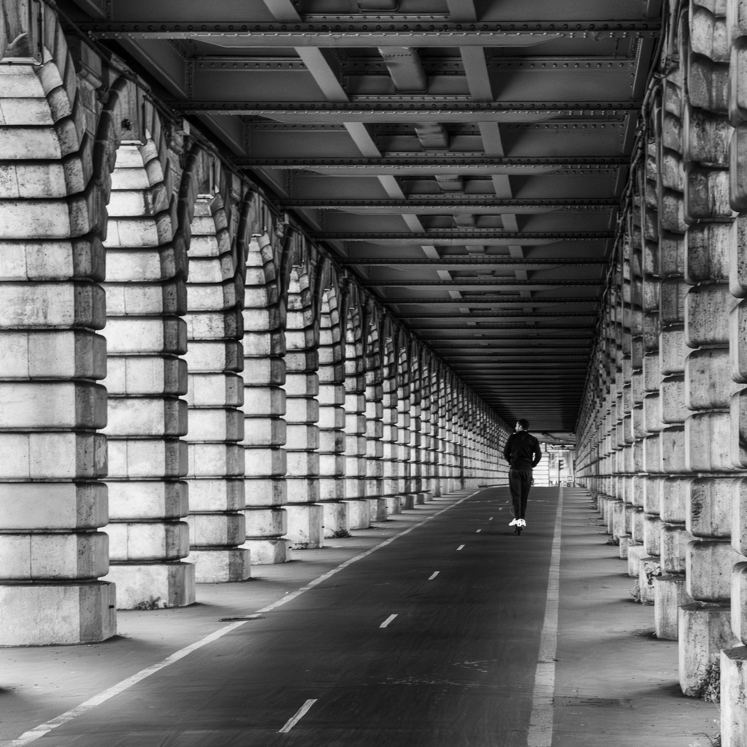Black and white photo of a lone person walking under a grand arched bridge colonnade with a vanishing point perspective.