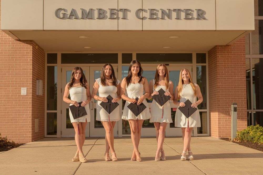 Five young women in white dresses holding graduation caps pose in front of the Gambet Center building.