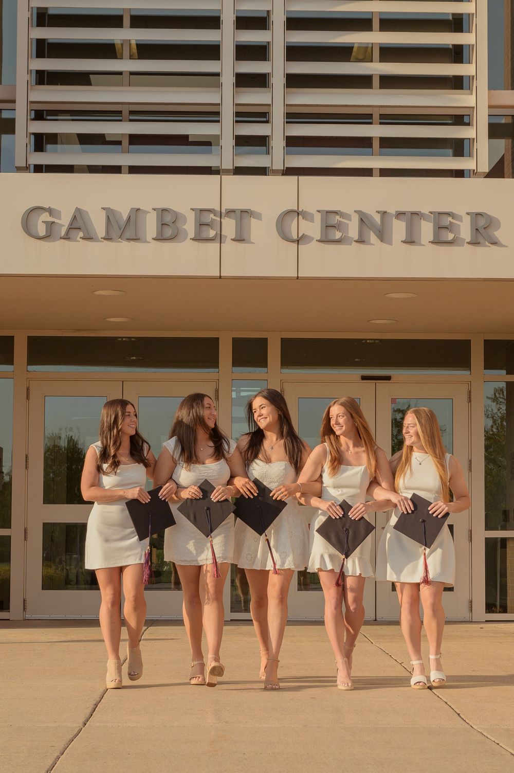 Five female graduates in white dresses hold caps outside the Gambet Center building.