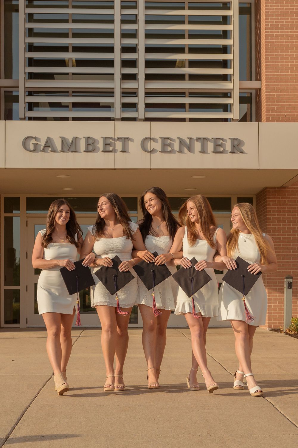 Five female graduates in white dresses hold caps outside DeSales University Gambet Center.
