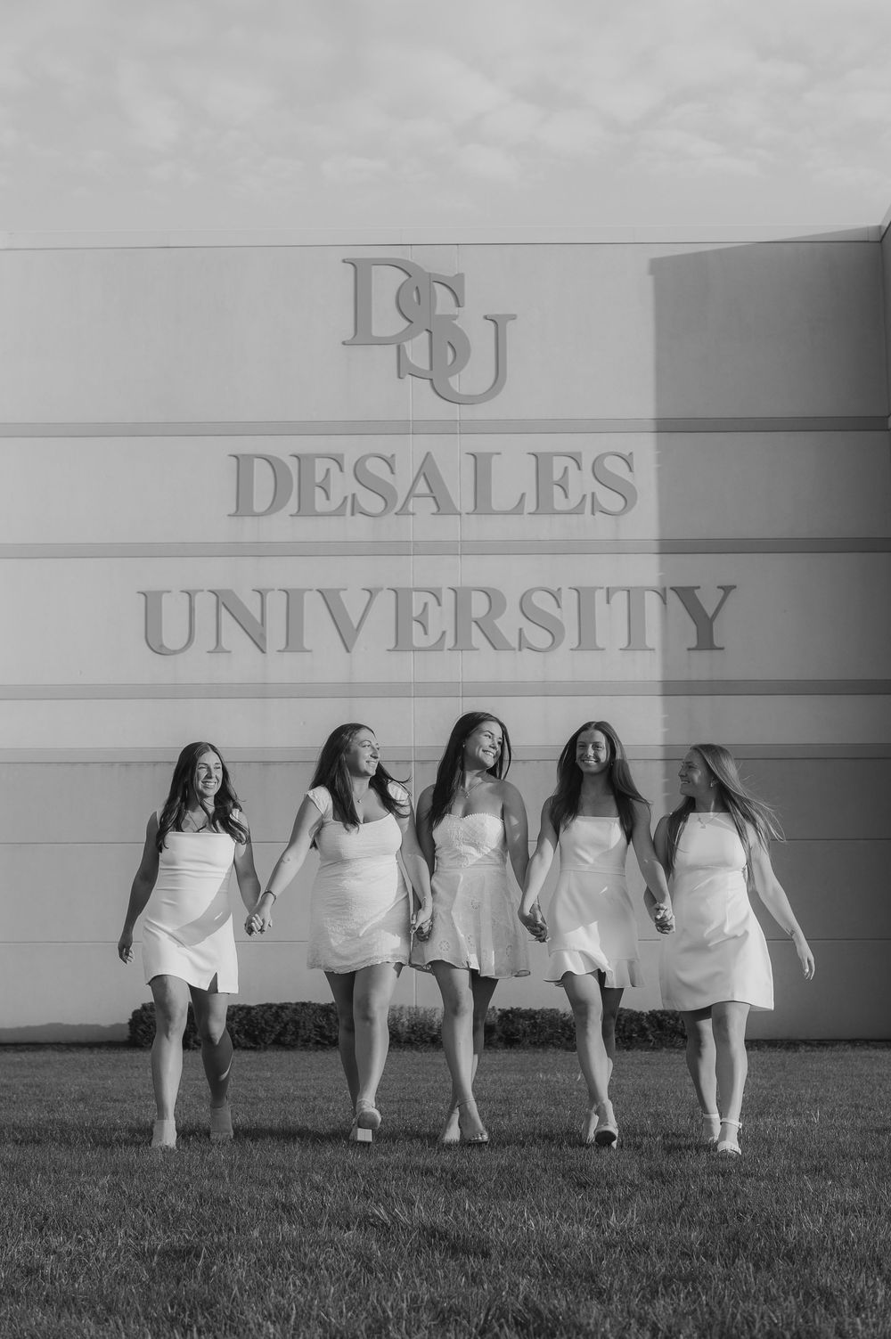 Five graduates in white dresses walk together in front of DeSales University sign.
