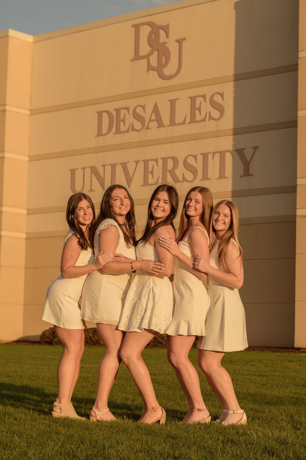 Five young women in white dresses pose together smiling in front of the DeSales University sign on a sunny day.