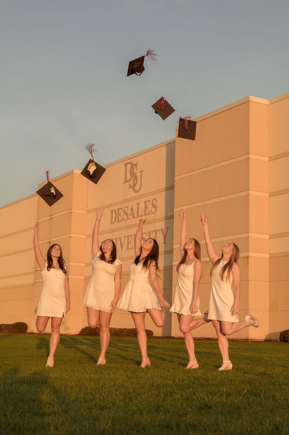 Five female graduates in white dresses toss caps in the air at DeSales University.
