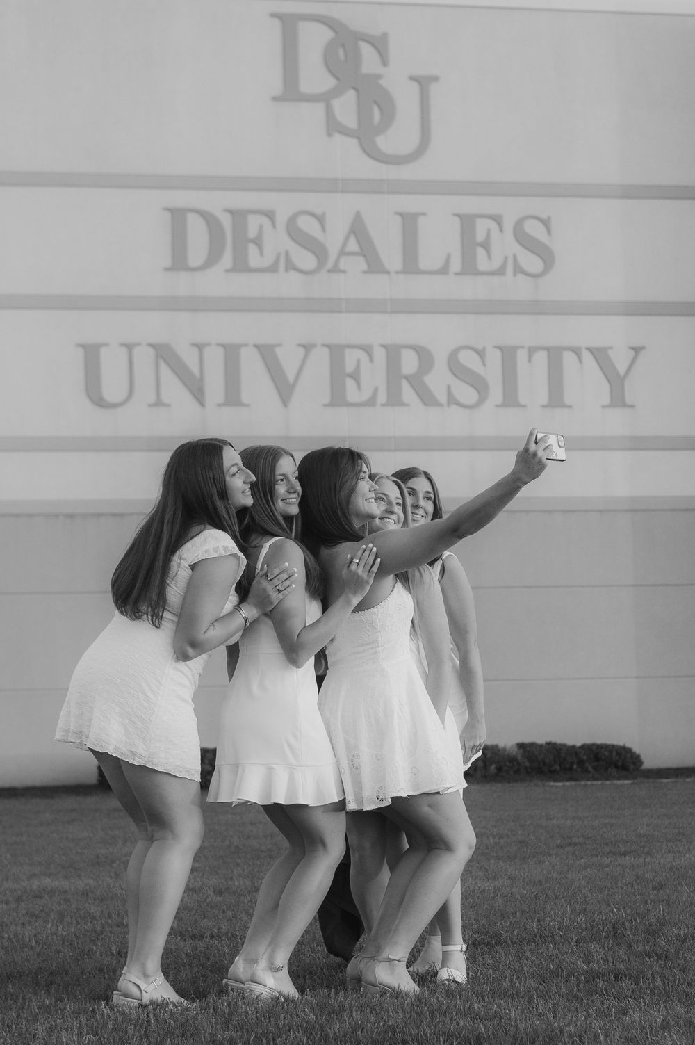 Group of young women in white dresses taking a selfie in front of DeSales University sign on campus.