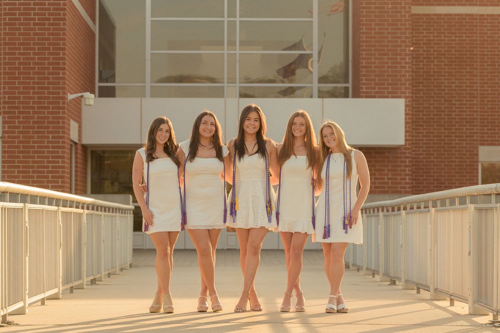 Five female graduates in white dresses and purple stoles pose on a bridge at golden hour.