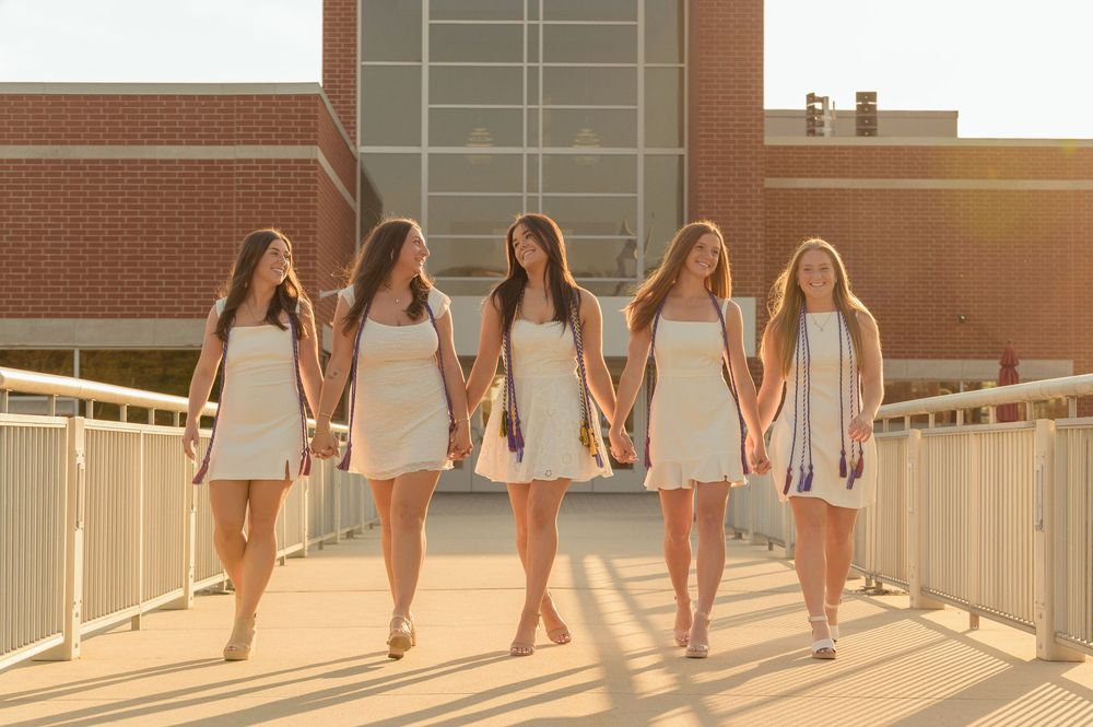 Five young women in white dresses walk hand-in-hand on a sunlit bridge near a brick building.