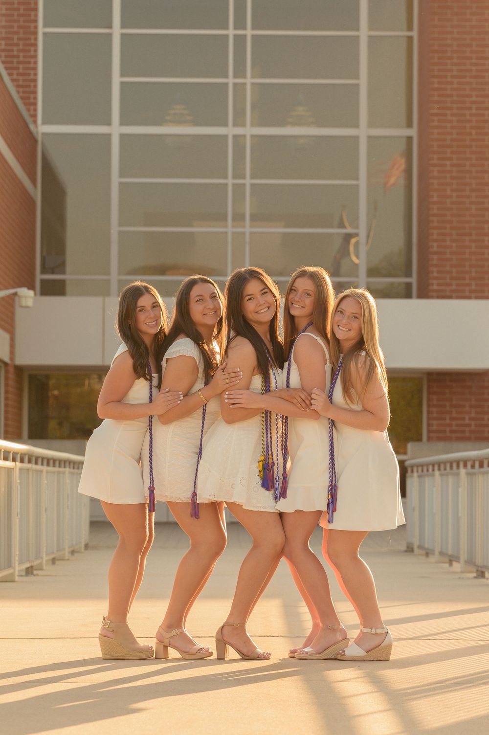 Five female graduates in white dresses pose together outside DeSales University building.