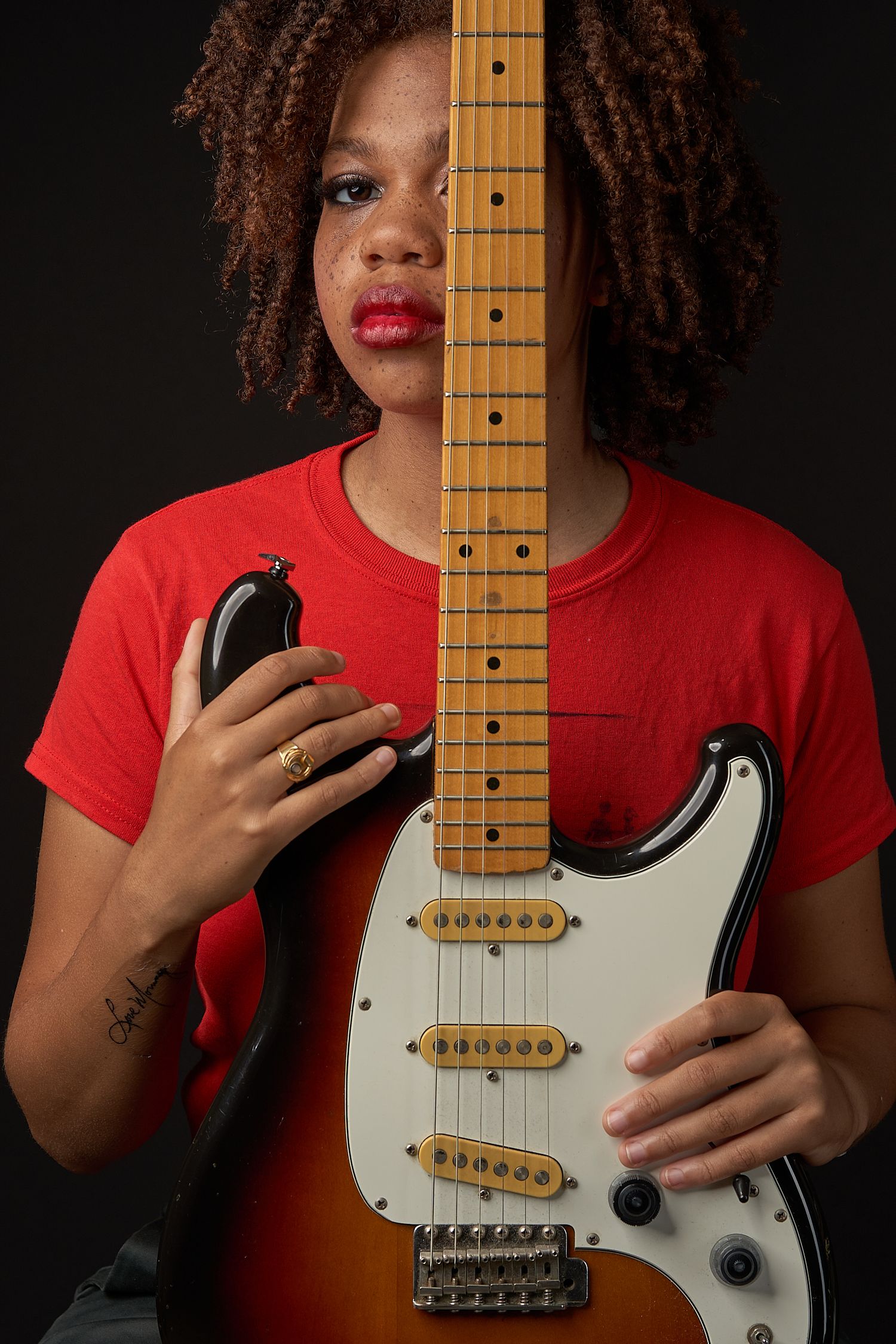 Young woman in red t-shirt holding electric guitar neck covering half her face against dark background.