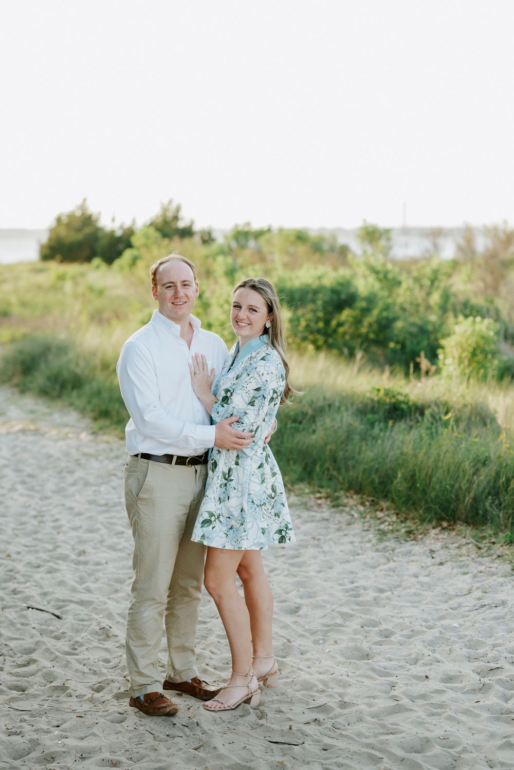 Couple embracing on sandy beach path, woman in floral dress and man in white shirt, with green dunes and water in background.