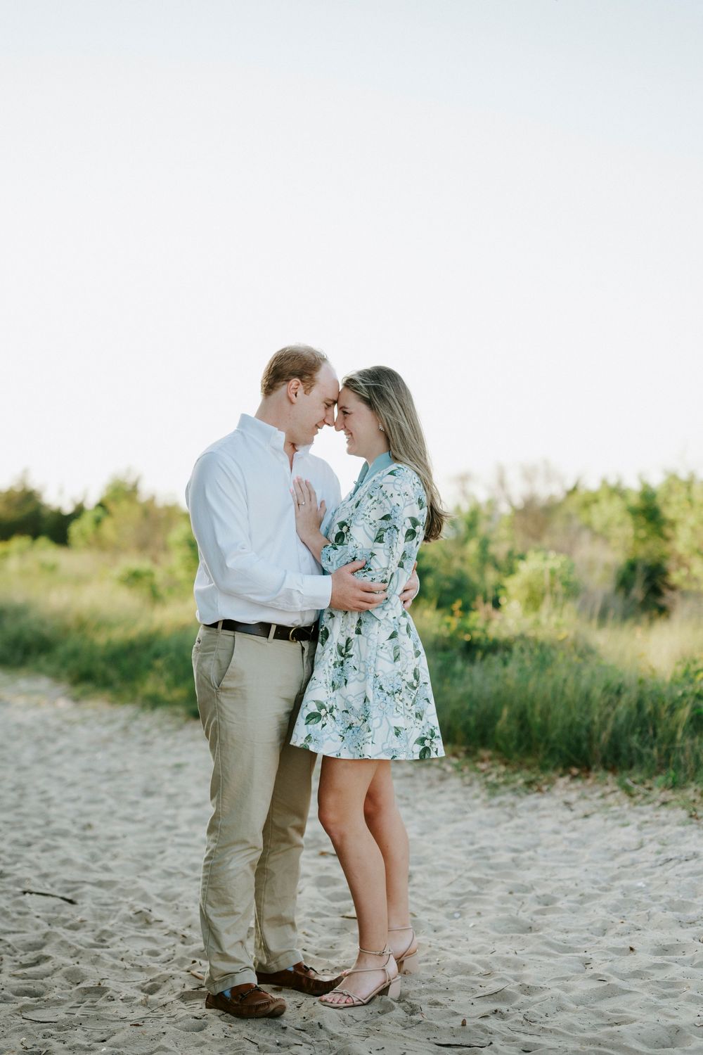 Couple in romantic embrace on sandy beach path, woman in floral dress, man in white shirt, surrounded by lush greenery.