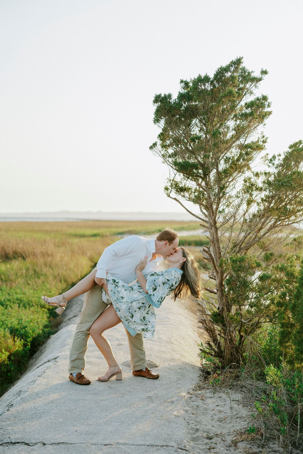 Couple sharing a romantic dip kiss on a sandy path surrounded by golden marsh grass and a pine tree at sunset.