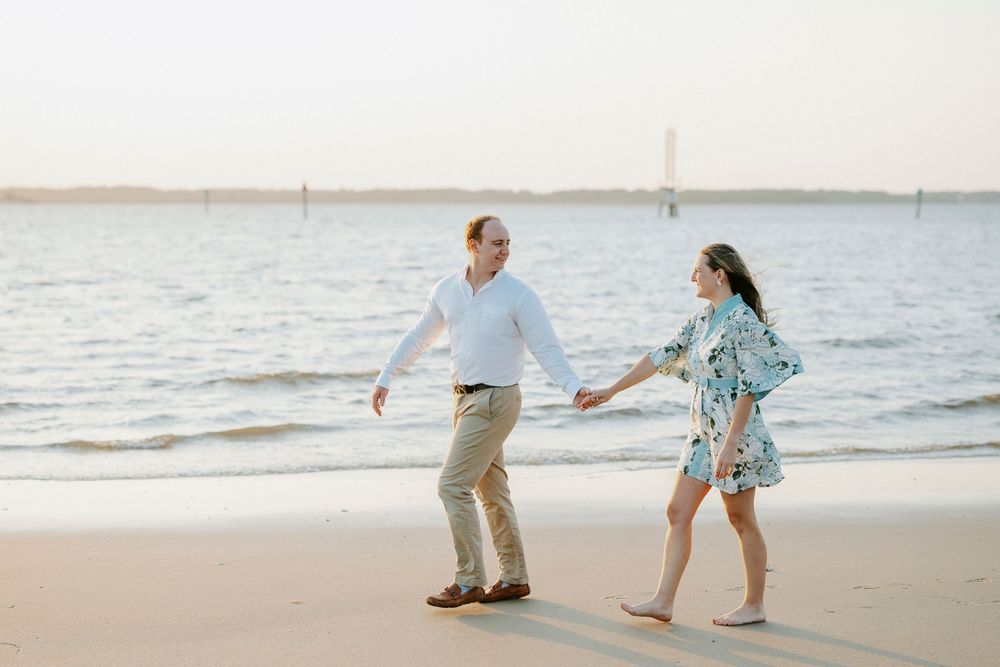 Couple holding hands walking on sandy beach near calm water at sunset in casual summer attire.